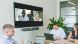 A photograph of a meeting space with a Logitech Rally Bar Mini mounted below a TV screen on the wall. There are three people sitting around a table.