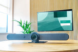 An image of the Logitech Rally Bar on a table in a meeting room. Behind the Rally Bar, there is a TV screen mounted on the wall showing the Google Meet home screen.