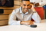 An image of a smiling man looking at the Series One Mic Pod in a meeting room. He has one hand on the table, next to the mic pod, and the other hand close to his face.